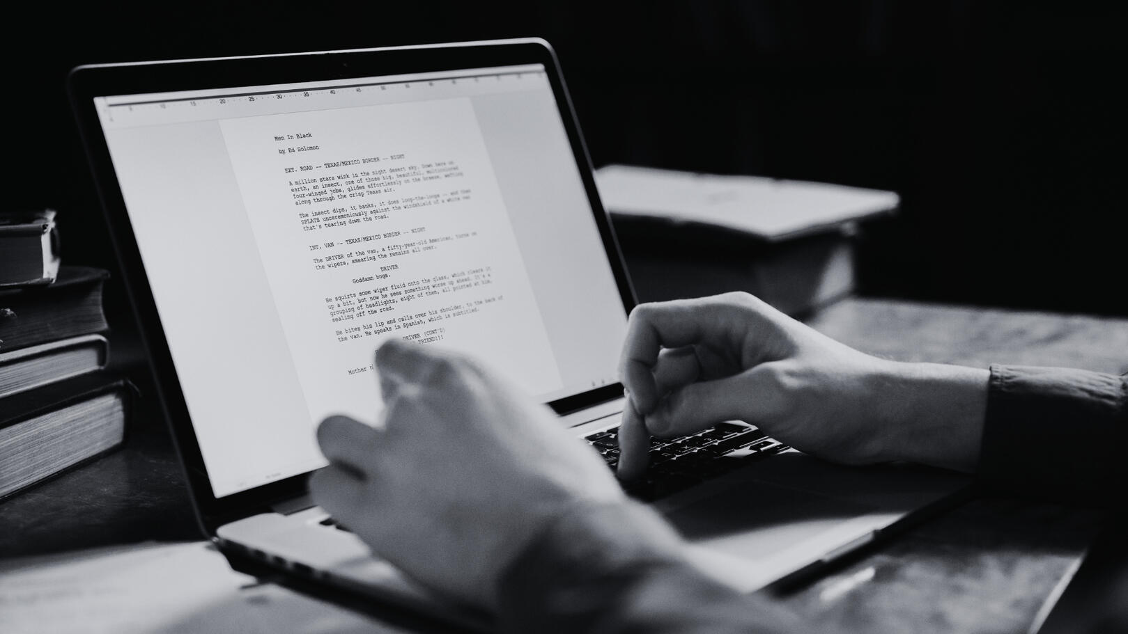Primo piano in bianco e nero di mani che scrivono una sceneggiatura tecnica su un laptop, con lo schermo acceso e dettagli di libri sullo sfondo sfocato.