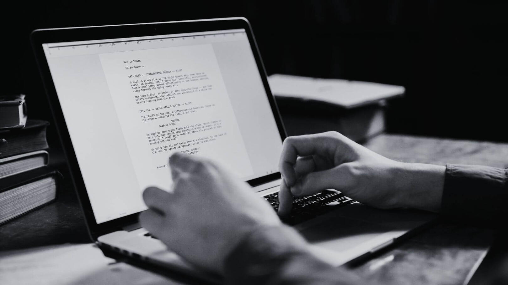 Screenwriting & Concept Development - Jessica Lima Primo piano in bianco e nero di mani che scrivono una sceneggiatura tecnica su un laptop, con lo schermo acceso e dettagli di libri sullo sfondo sfocato.