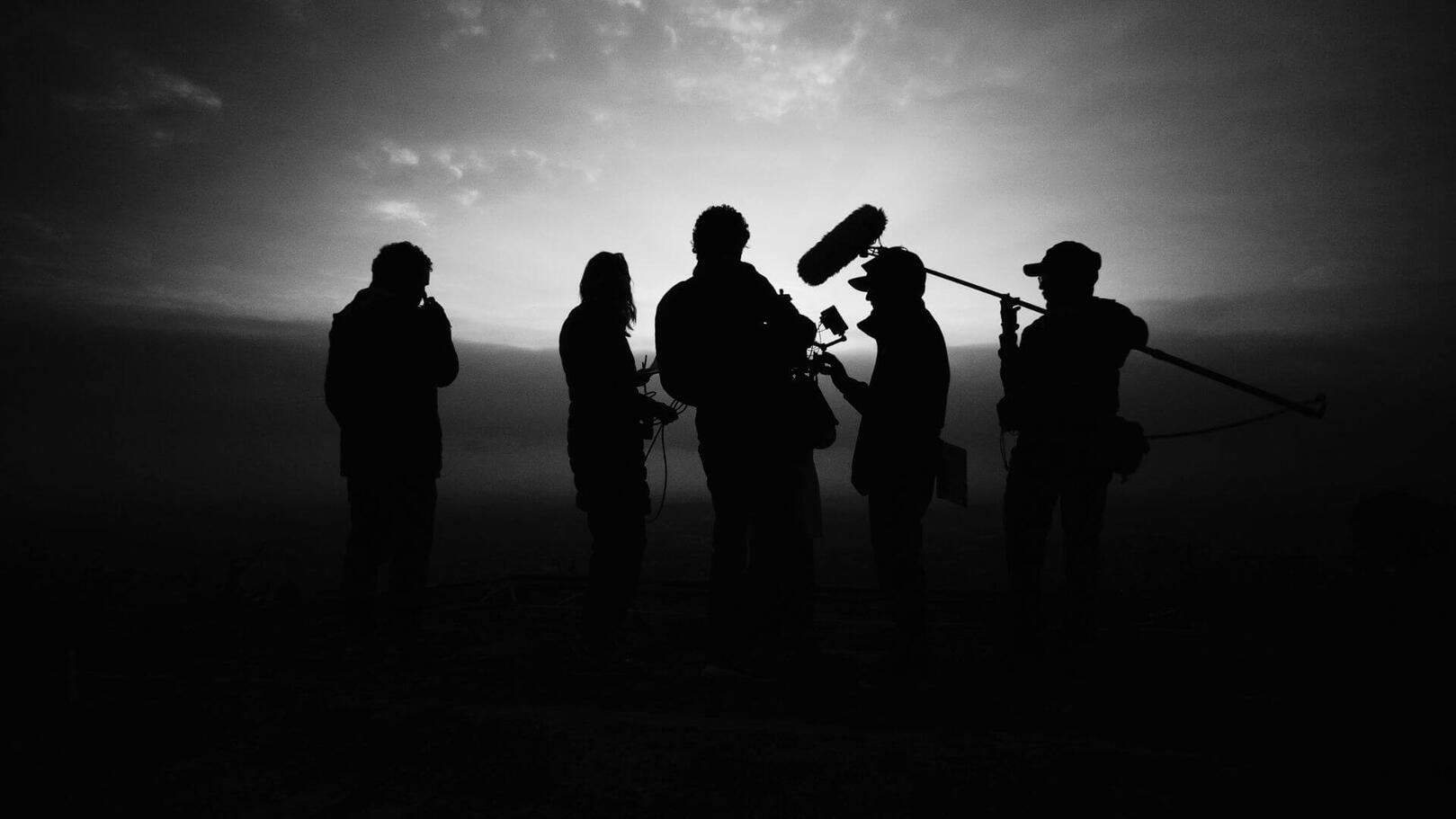 Leadership e Direzione di Troupe — Coordinamento Audiovisivo | Jessica Lima Un gruppo di lavoro della troupe cinematografica concentrato a esaminare le riprese al monitor sotto fasci di luce da set.