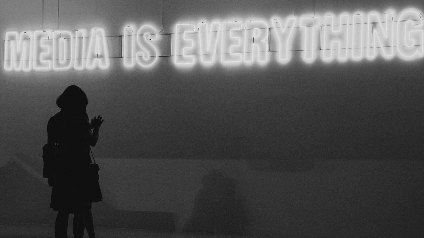 Promotion & Media Planning — La Strategia Media e Diffusione dei Contenuti | Jessica Lima Una silhouette di persona di spalle osserva una scritta al neon luminosa che recita: "MEDIA IS EVERYTHING".